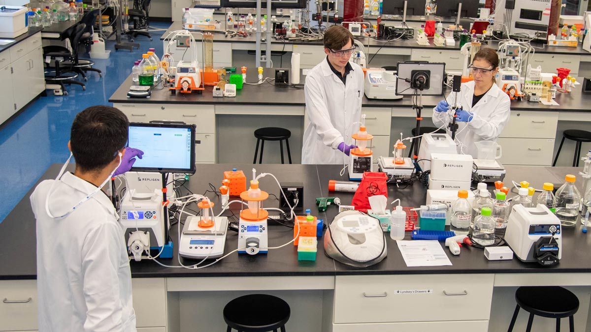 Three people work in a lab environment with many pieces of equipment on multiple work stations