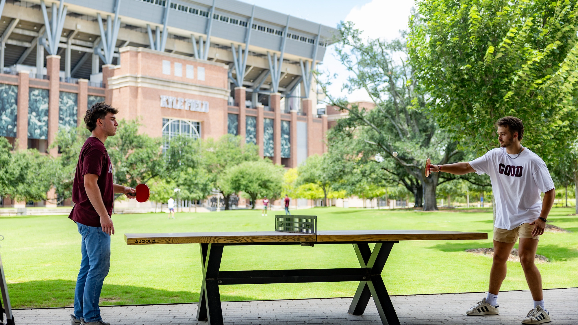 Two young men are playing a game of ping pong outdoors, with a large sports stadium visible in the background.