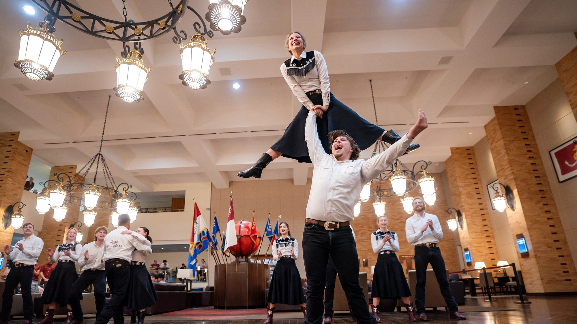 A couple performs a lively dance with the woman being lifted by her partner in a festive setting, surrounded by other dancers in traditional attire.