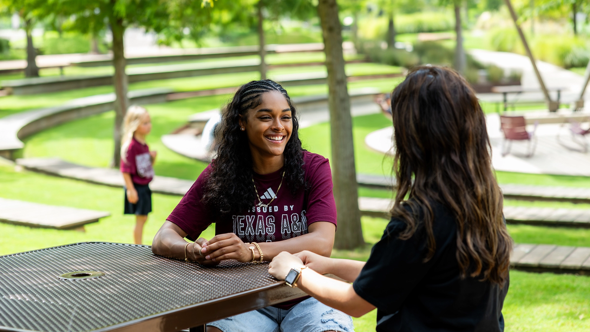 Two young women engage in conversation at an outdoor table, enjoying a sunny day in a park-like setting. The background features grassy areas and seating arrangements.