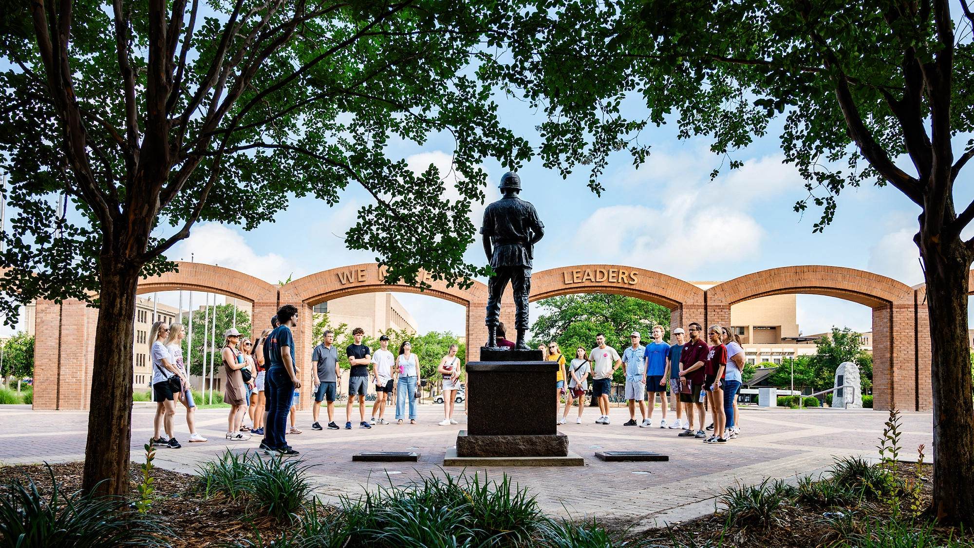 A group of people gathers around a statue at a monument under trees, with the words "WE MAKE LEADERS" prominently displayed in the background.
